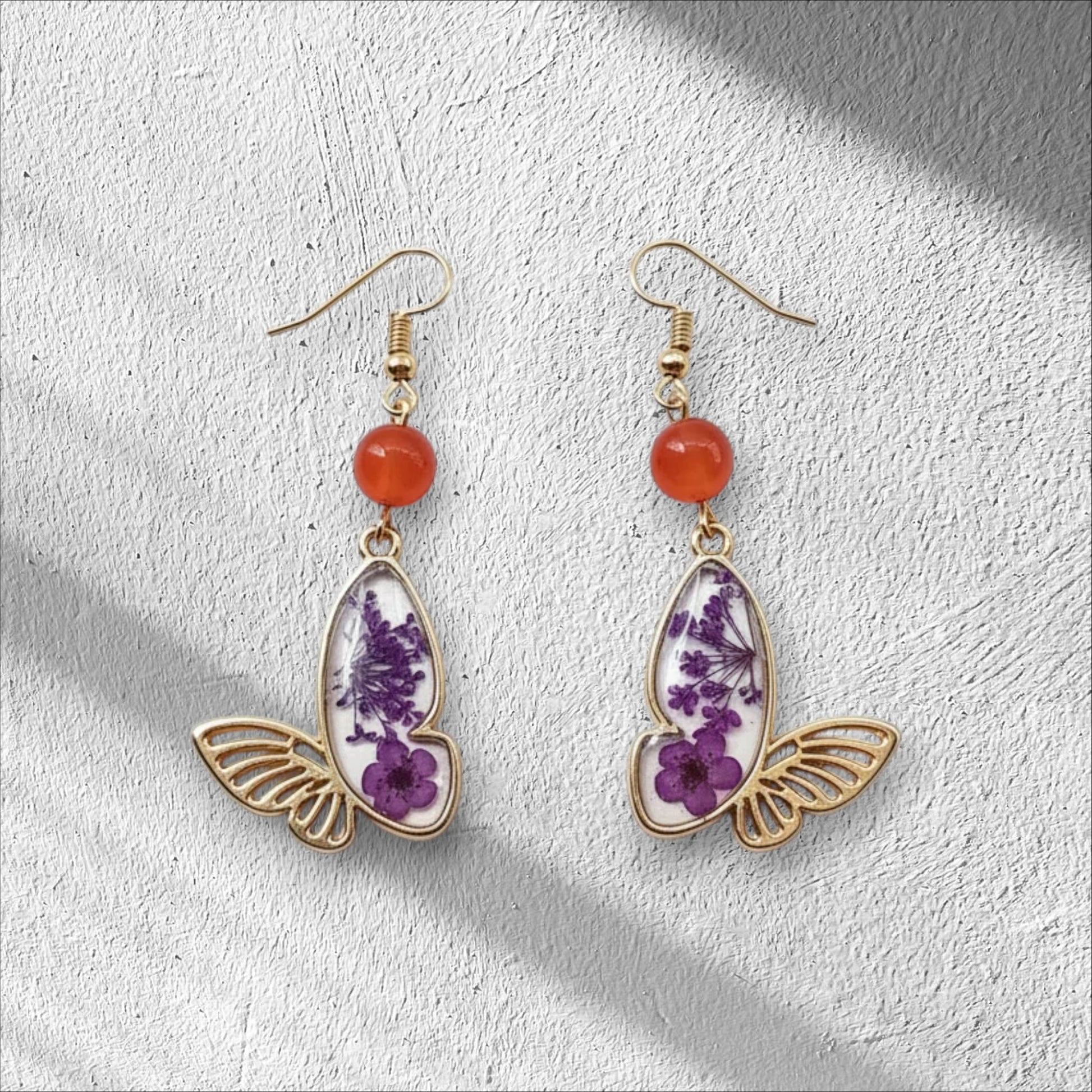 Alyssa's House gold-tone butterfly earrings featuring purple pressed flowers and red bead accents.